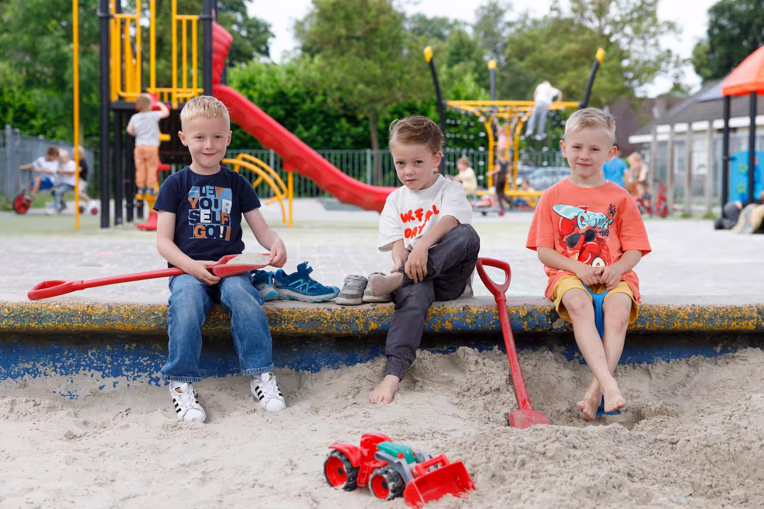 Drie jongens zitten in een zandbak bij een speeltuin met rode en blauwe speeltuinattributen.