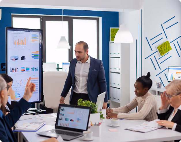Business team in meeting room reviewing charts on large screen and laptops during discussion.
