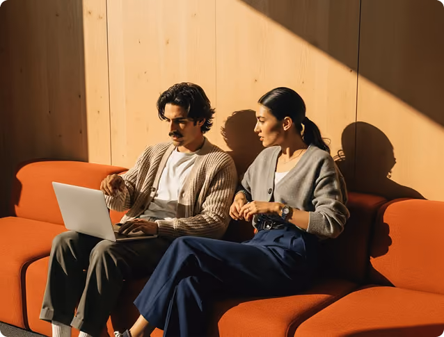 Man and woman sitting on orange couch looking at a laptop in a sunlit room with wooden walls.