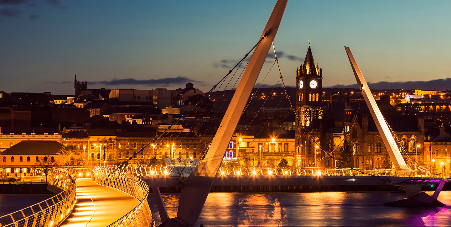 Illuminated modern pedestrian bridge over water with historic clock tower and buildings lit at dusk.