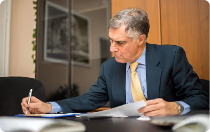 Man in suit and yellow tie writing while reviewing papers at a desk in an office.