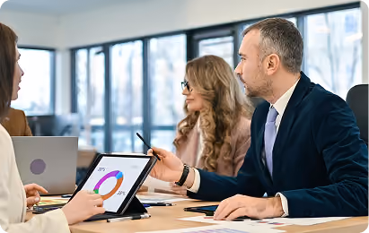 Three professionals discussing data with a tablet displaying a pie chart at a meeting table.