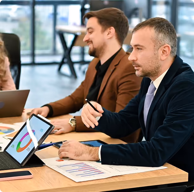 Two men at a table analyzing charts on a tablet and paper in a bright office setting.