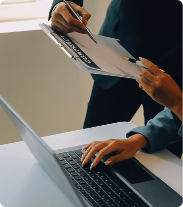 Person typing on a laptop while another points at a contract on a clipboard with a pen.