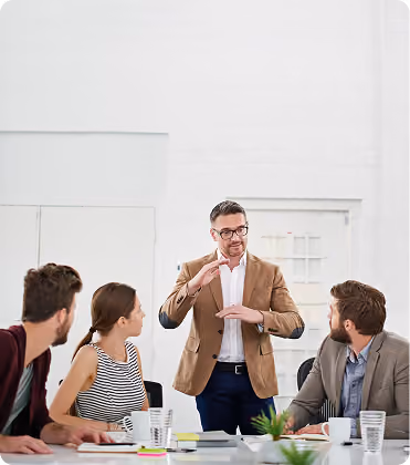 Man in brown blazer explaining with hand gestures to three seated colleagues in a meeting room.