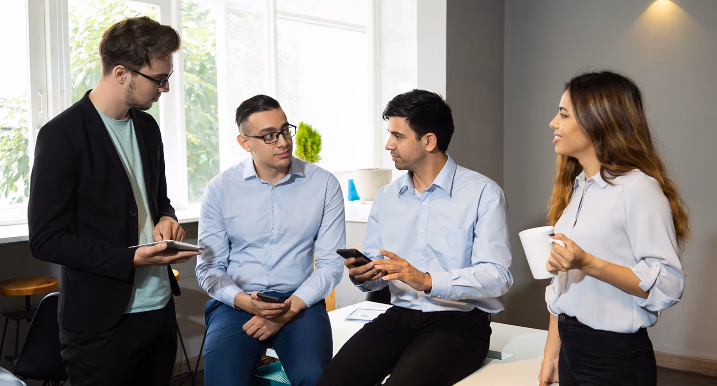 Four professionals in casual office wear having a discussion, two holding phones and one with a coffee mug.