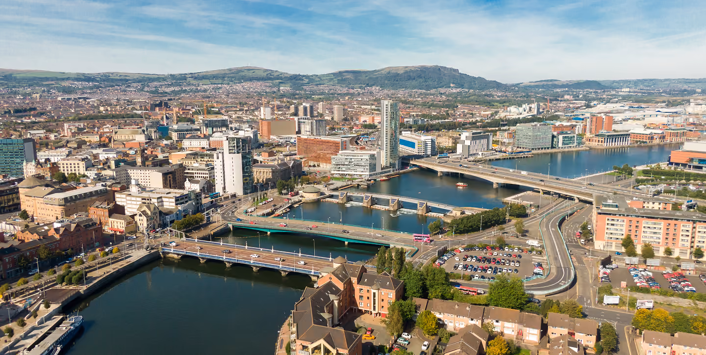 Aerial view of a city with bridges over a river, buildings, roads, parking lots, and hills in the background.