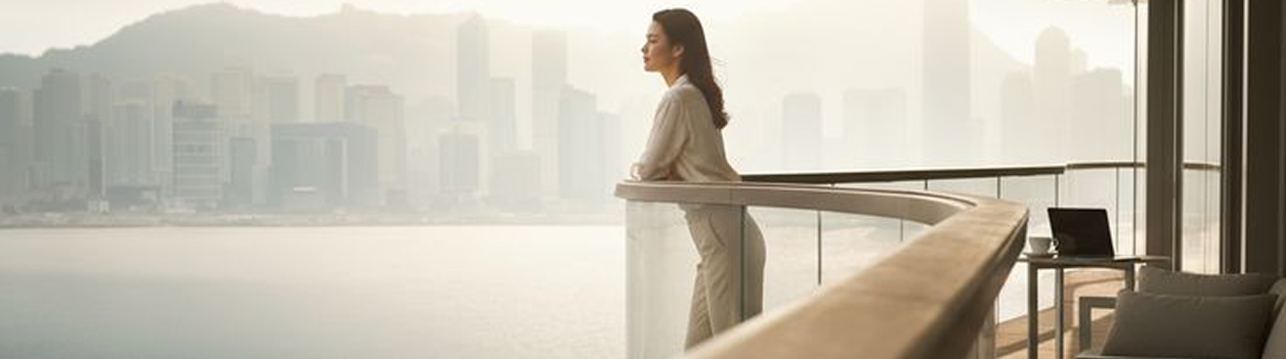 Woman in beige outfit leaning on curved balcony railing overlooking foggy city skyline across water.