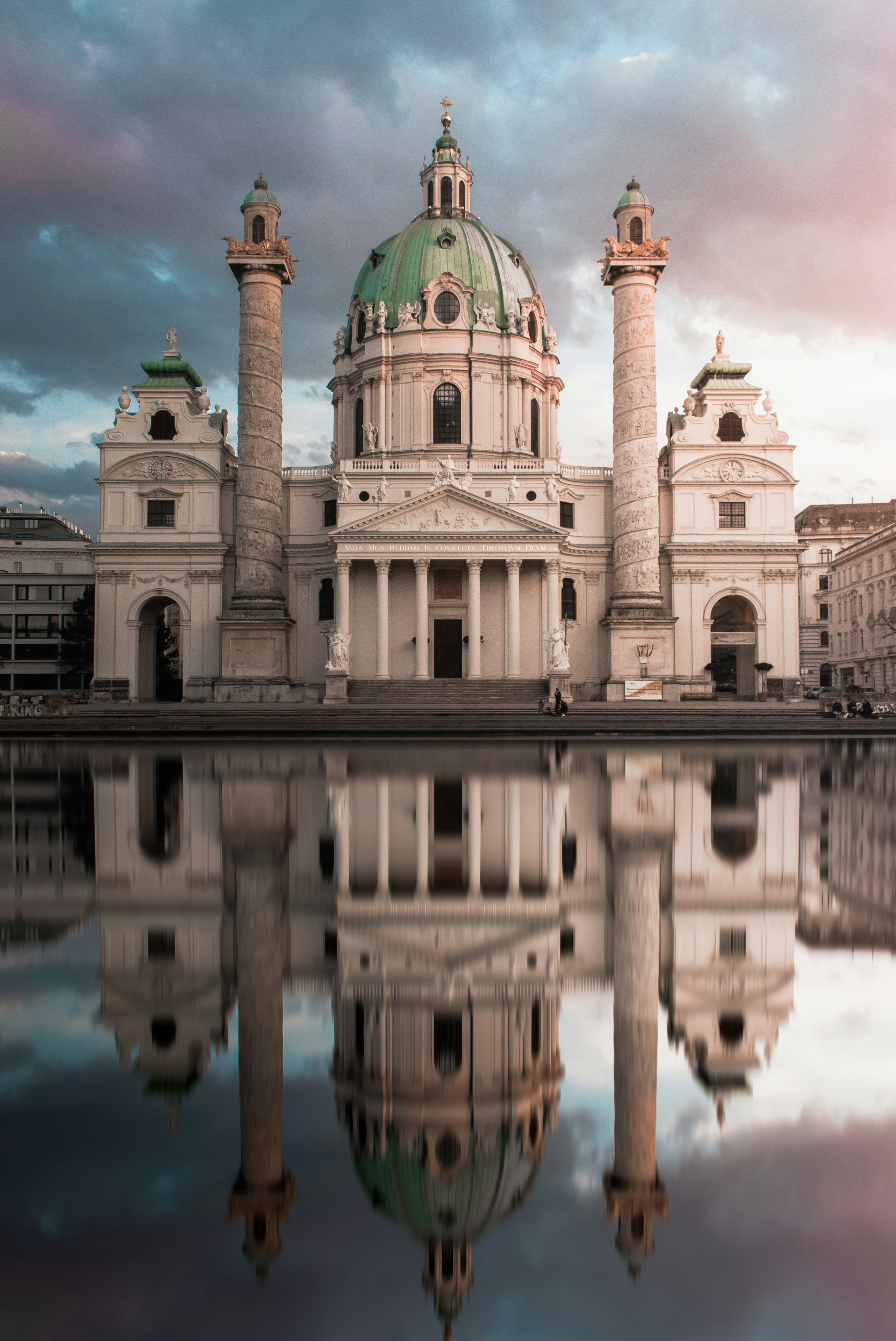 Baroque church with a green domed roof and two columns topped with golden sculptures, reflected in the water.