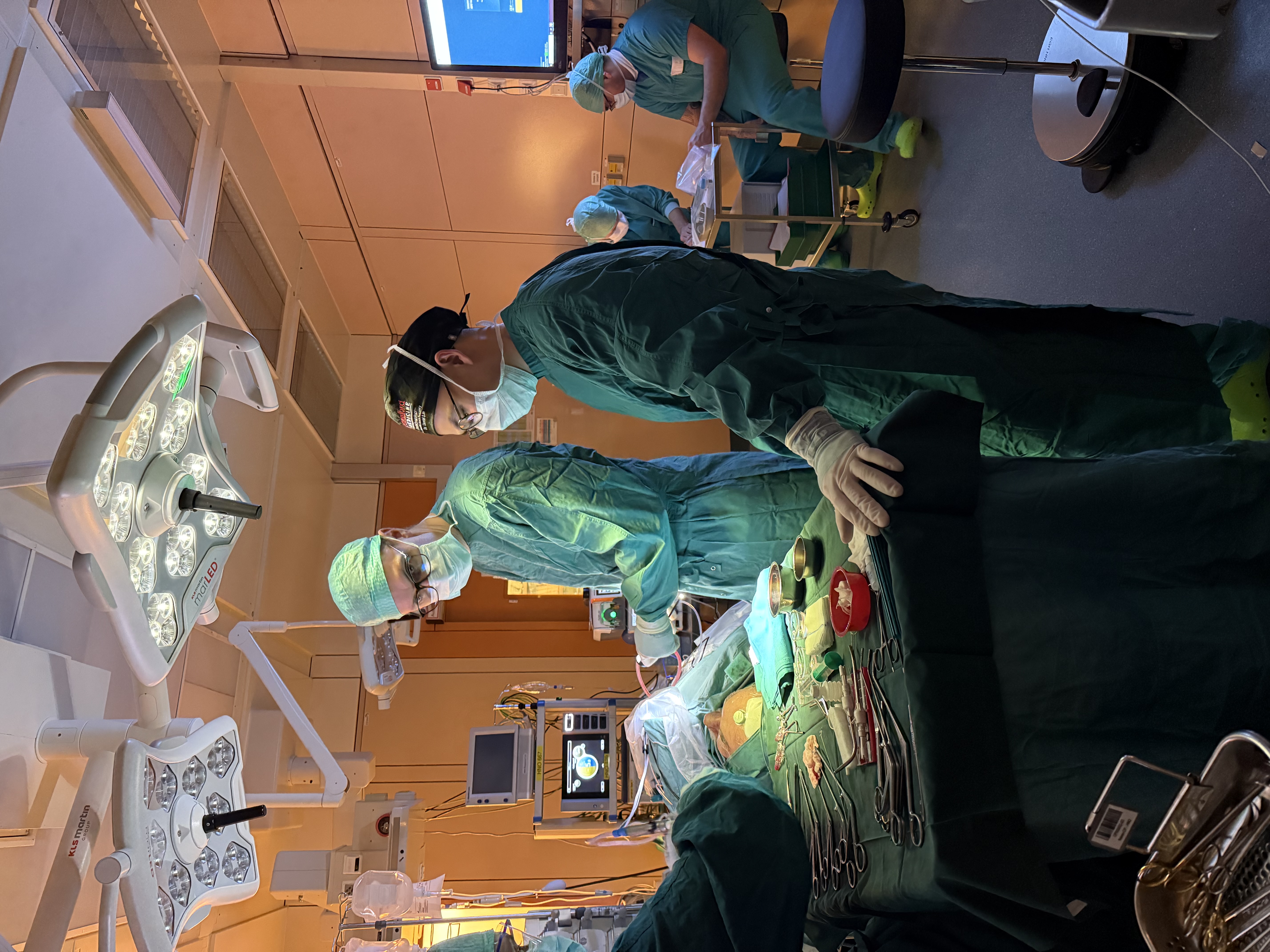 Surgeons in green scrubs performing an operation in an OR, with an instrument table in the foreground and monitors and staff in the background; a calendar overlay reads "24 October."