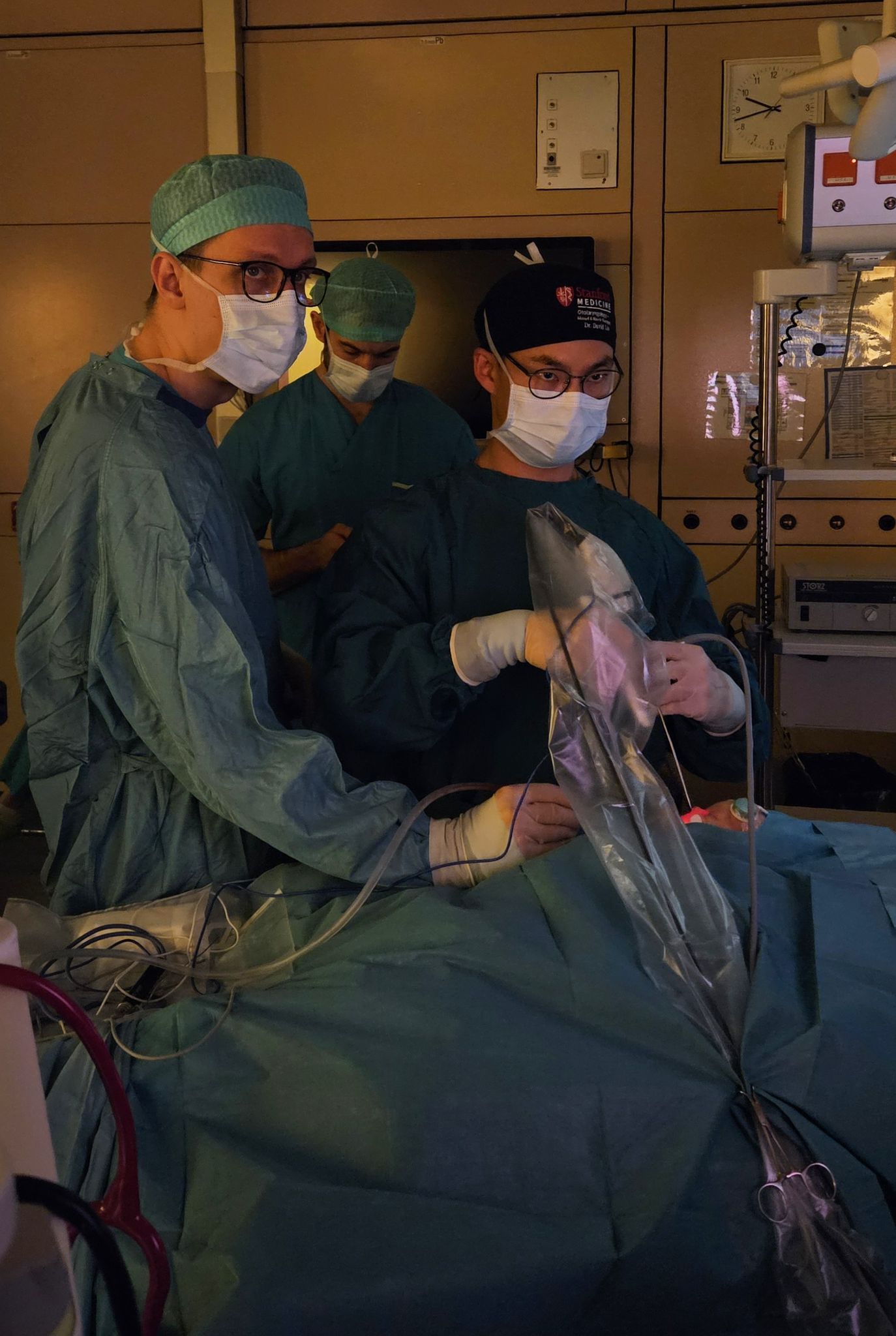 Three surgeons in scrubs and masks performing a procedure, holding a sterile drape over the patient with surgical equipment and monitors in the background.