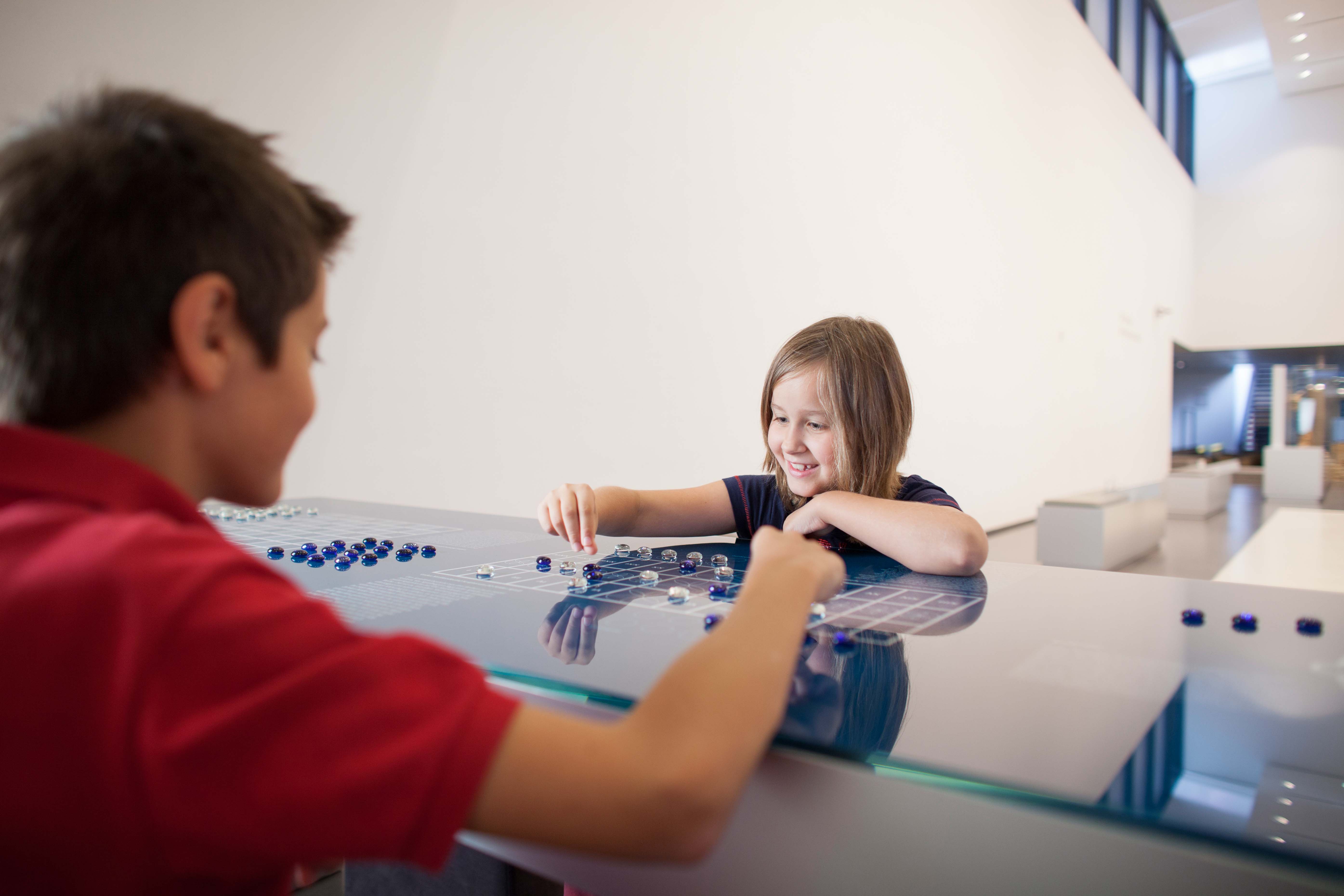 Zwei Kinder, ein Junge und ein Mädchen, spielen ein Spiel mit blauen und klaren Glassteinen auf einem gläsernen Spielbrett in einem modernen Innenraum.