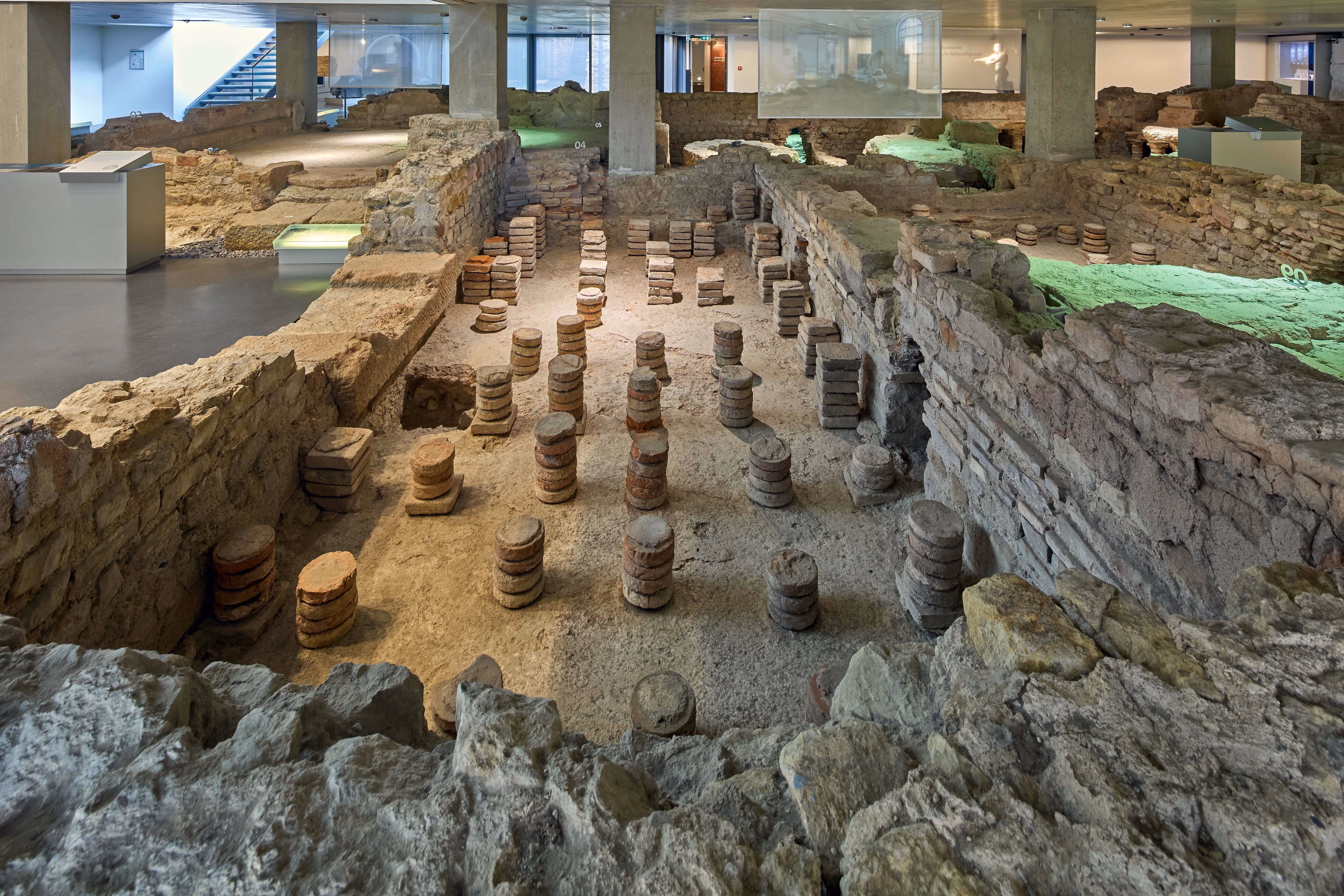 Blick auf die gut erhaltenen römischen Thermenruinen mit hypokaustischen Säulen im Boden zur Beheizung des Fußbodens, umgeben von Steinmauern in einem modernen Museum.