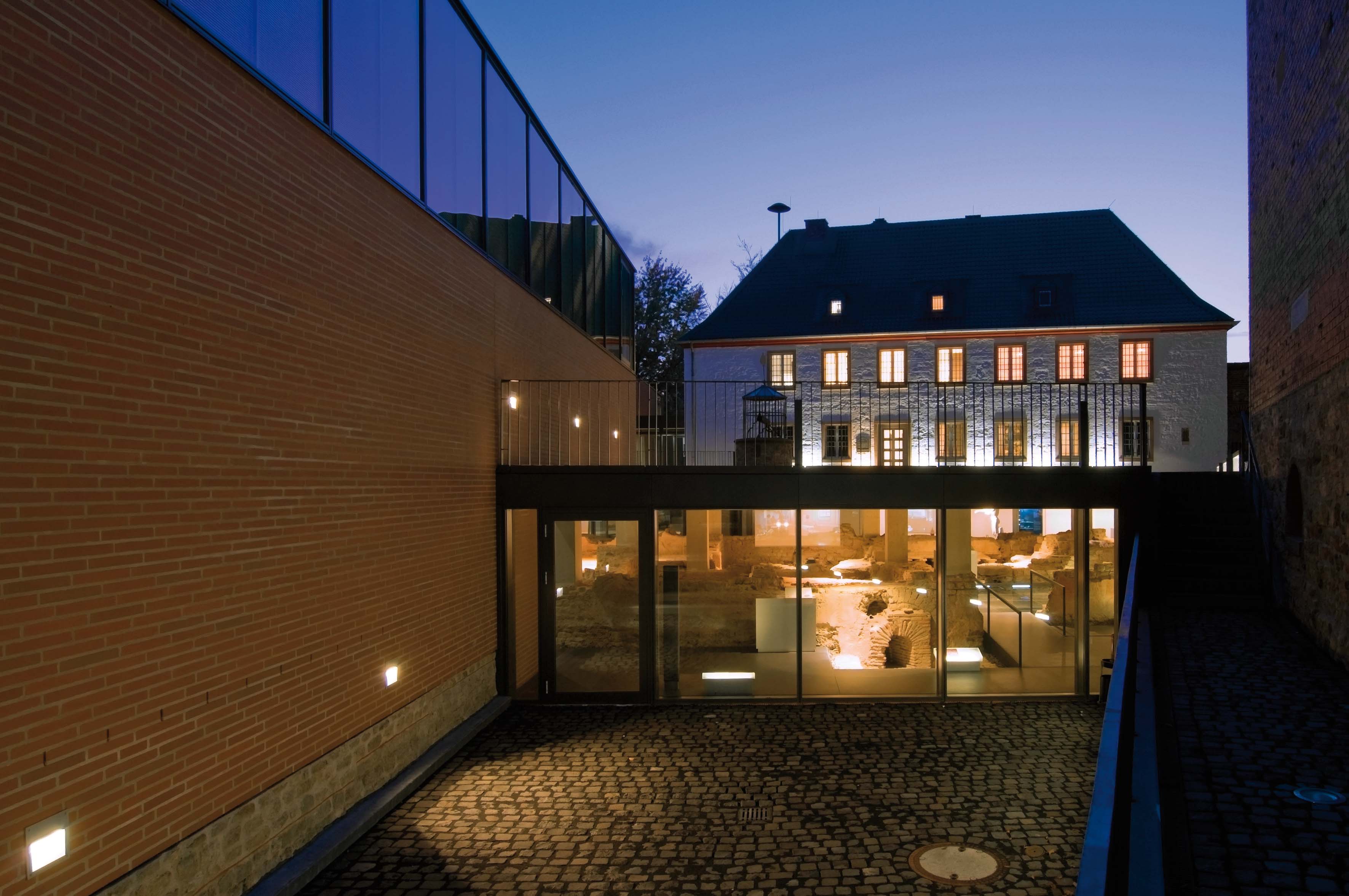 Nachtszenario eines Gebäudekomplexes mit moderner Glasfassade im Vordergrund, beleuchteten archäologischen Ruinen im unteren Bereich und einem historischen, weiß getünchten Haus mit leuchtenden Fenstern im Hintergrund.