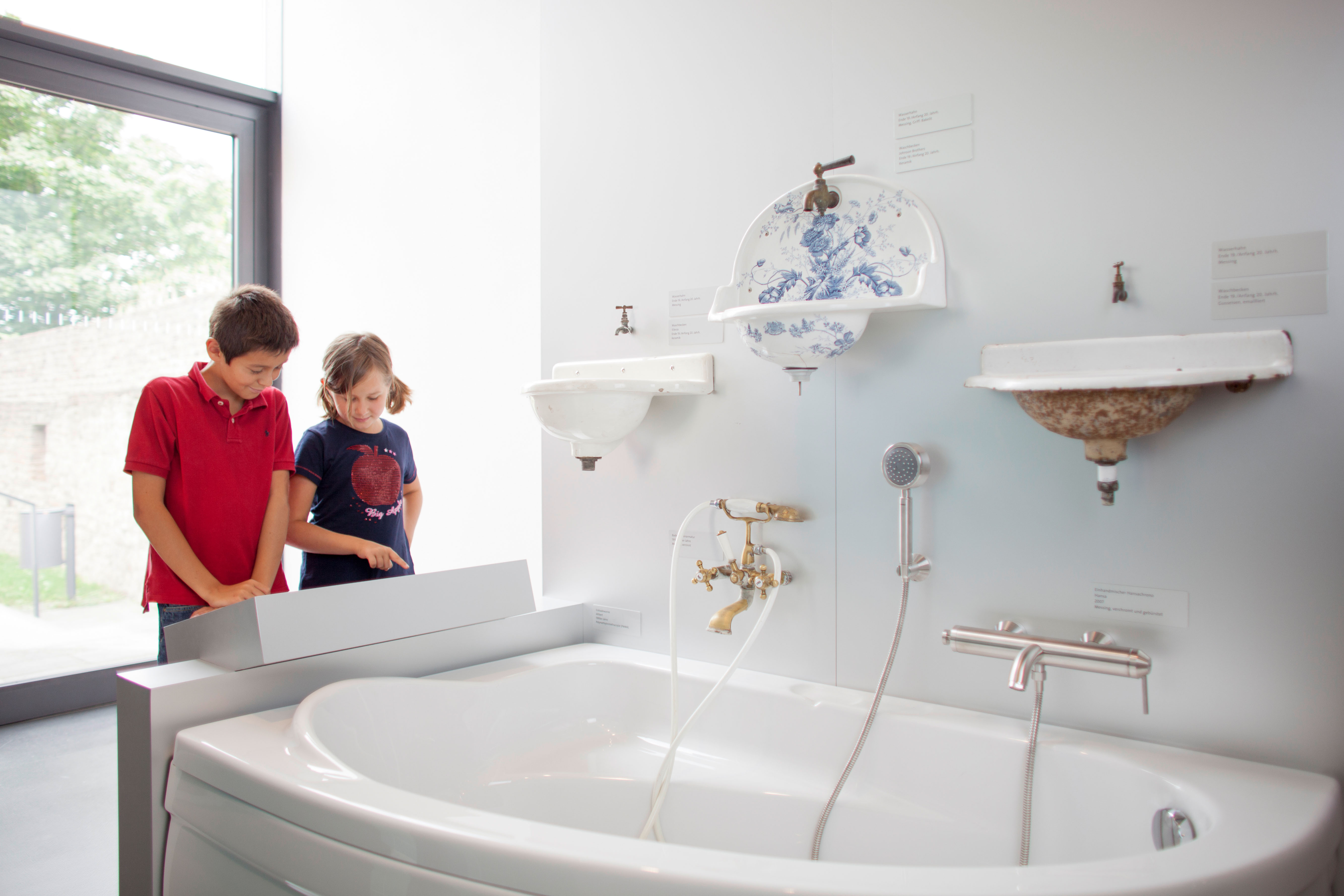 Zwei Kinder, ein Junge in rotem Polohemd und ein Mädchen in blauem T-Shirt mit Apfelmotiv, betrachten eine Ausstellung von historischen Waschbecken und Badewanne in einem hellen Raum mit großem Fenster.