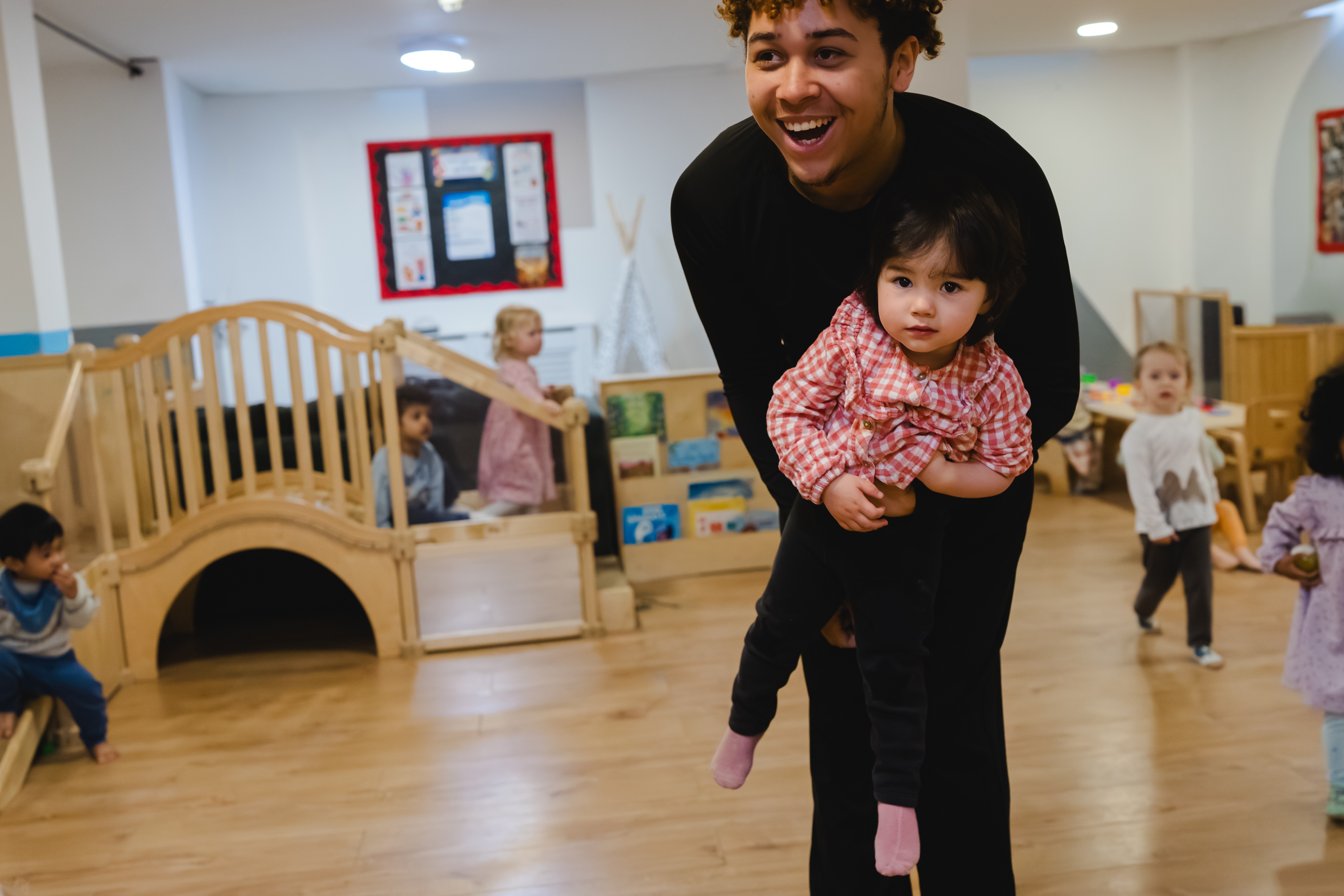 Settling in. A nursery practitioner carries a smiling toddler through the playroom while other children play nearby