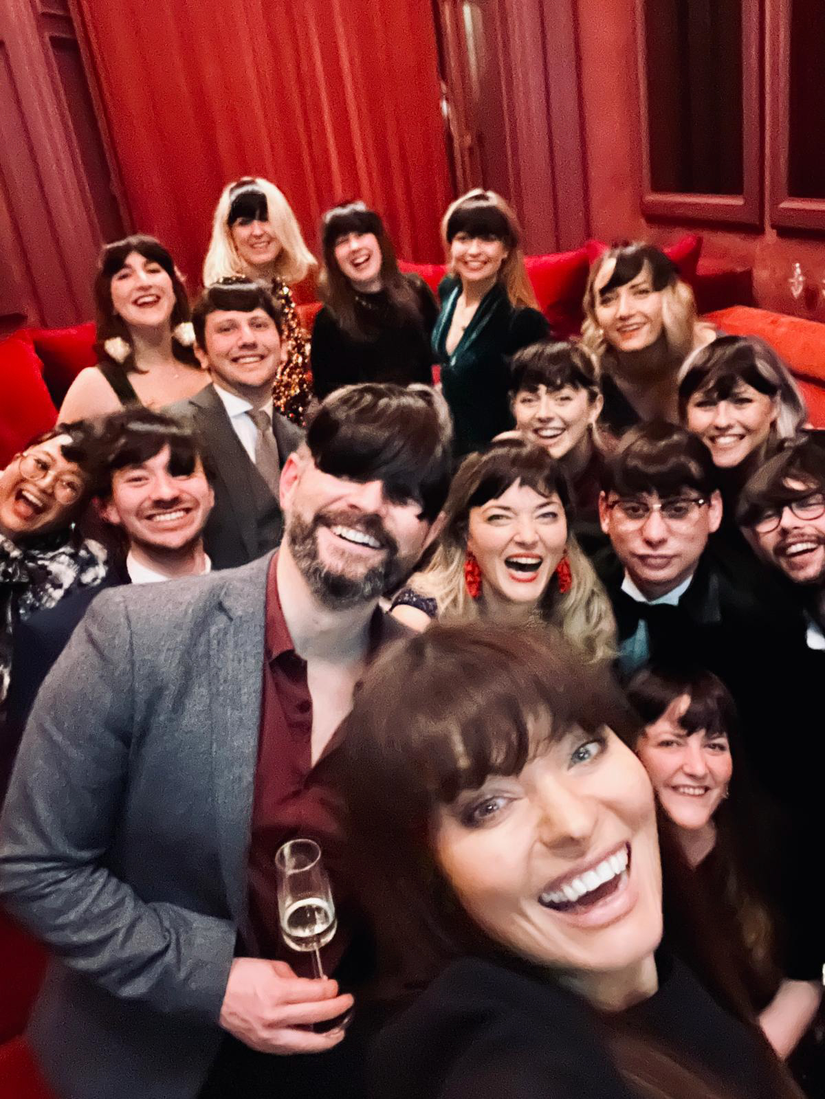 Group selfie of joyful adults dressed formally in a room with red curtains and couches.