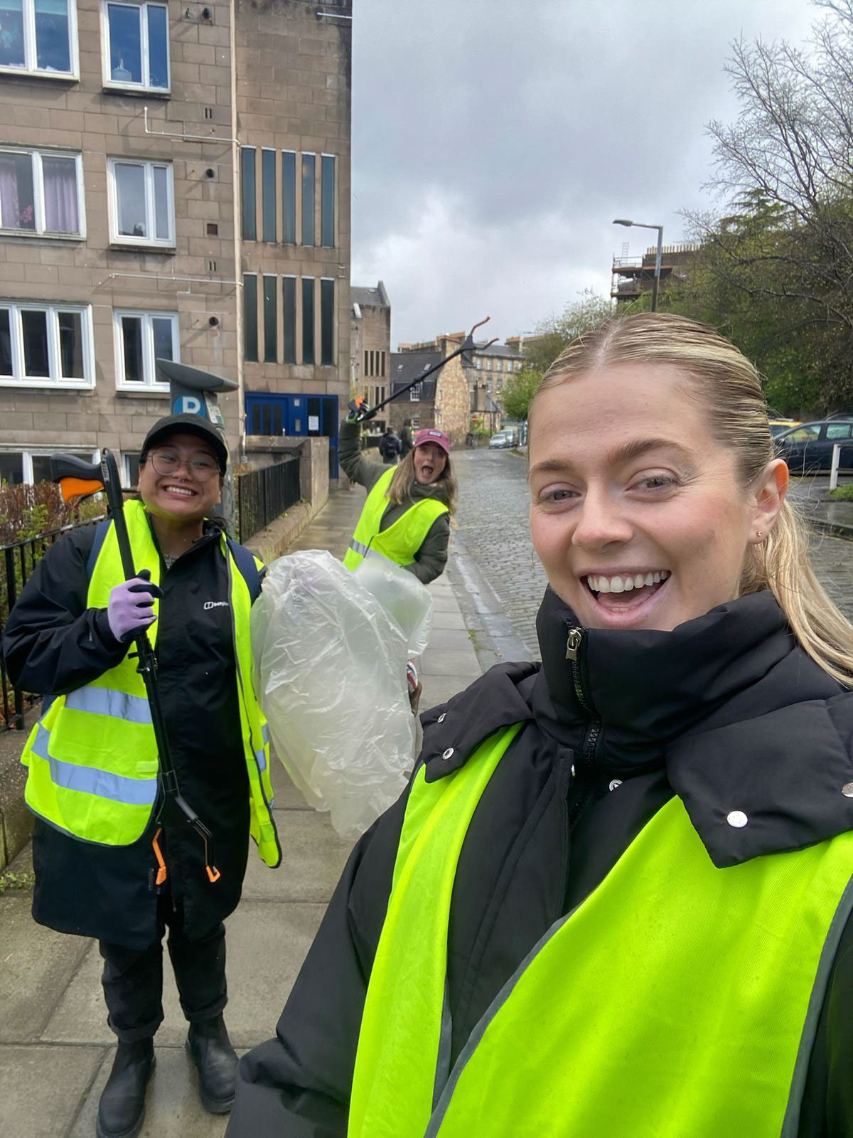 Three women in yellow vests smiling and holding litter-picking tools on a sidewalk by buildings.
