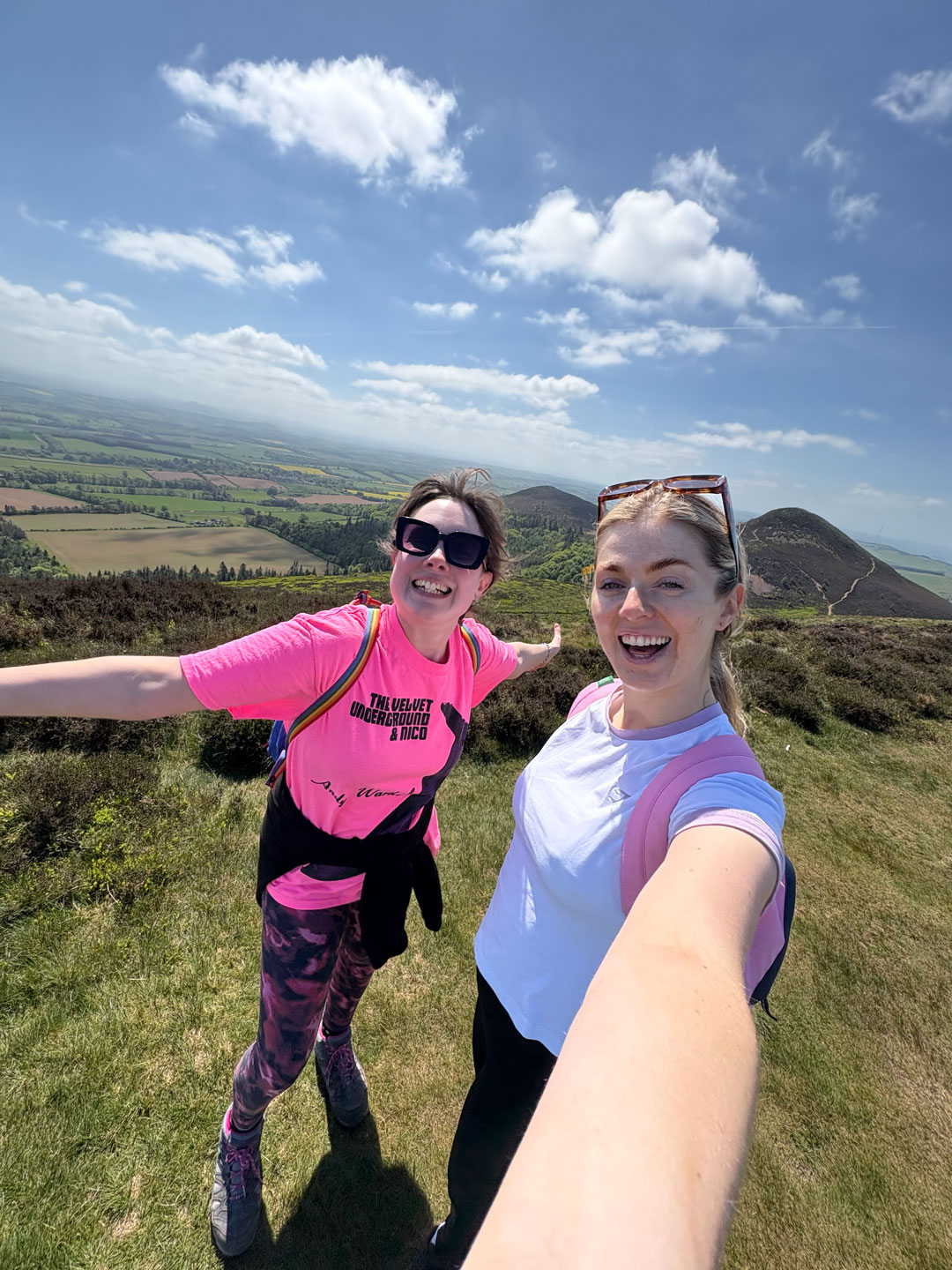 Two women smiling and taking a selfie on a sunny hill with green fields and blue sky behind them.