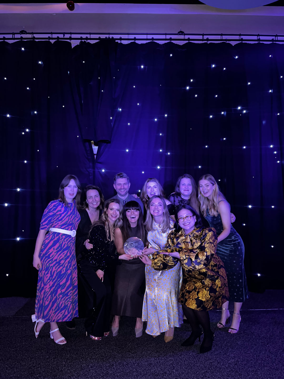 Group of nine people posing with an award in front of a starry curtain backdrop at an event.
