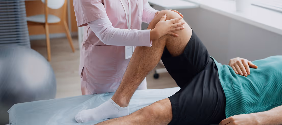 Physical therapist in pink scrubs assisting a patient by bending his knee on an examination table.
