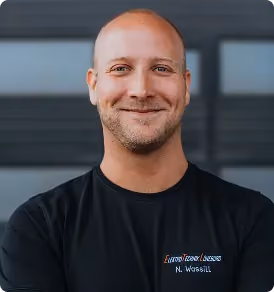 Lächelnder Mann mit kurzem Haar und schwarzem Shirt vor einer dunklen, unscharfen Wand.