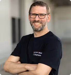 Mann mit Brille, Bart und schwarzem T-Shirt lächelt mit verschränkten Armen vor unscharfem Hintergrund.