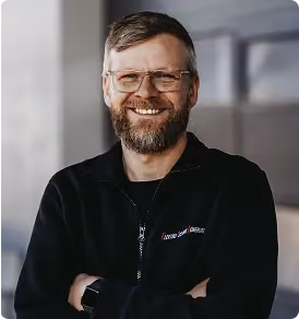 Mann mit Brille und Bart, lächelnd, trägt schwarzen Pullover, steht mit verschränkten Armen vor unscharfem Hintergrund.