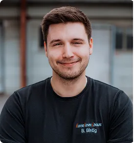 Lächelnder Mann mit kurzen braunen Haaren trägt ein schwarzes T-Shirt vor Gebäude.