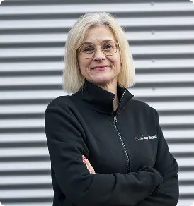 Lächelnde Frau mit blonden Haaren, Brille und schwarzem Oberteil vor grauem gestreiftem Hintergrund.