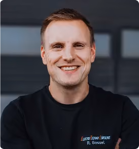 Porträt eines lächelnden Mannes mit kurzem Haar in schwarzem T-Shirt vor dunklem Hintergrund.
