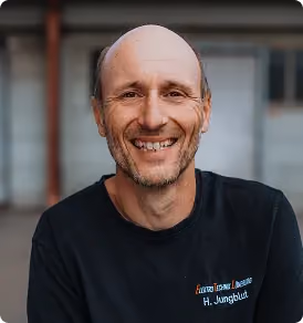 Lächelnder Mann mit Glatze und schwarzem T-Shirt vor unscharfem Hintergrund.