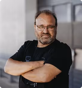 Mann mit Brille, Bart und schwarzem Shirt steht vor einem grauen Hintergrund mit verschränkten Armen.