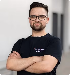 Junger Mann mit Brille und Bart, verschränkte Arme, lächelnd vor unscharfem Hintergrund.