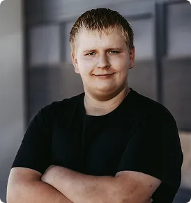 Junger Mann mit blonden Haaren und schwarzem T-Shirt, verschränkte Arme, leicht lächelnd.