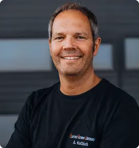 Ein lächelnder Mann mit dunklem T-Shirt vor unscharfem, dunklem Hintergrund.