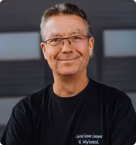 Lächelnder Mann mit Brille und dunklem T-Shirt vor unscharfem Hintergrund.