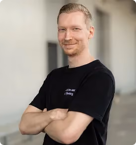 Junger Mann mit verschränkten Armen lächelt vor unscharfem urbanem Hintergrund.