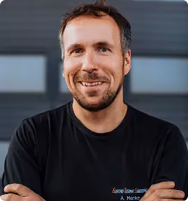 Lächelnder Mann mit kurzem Bart und schwarzen T-Shirt vor einer dunklen vertäfelten Wand.