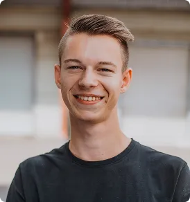 Junger Mann mit kurzem Haar lächelt vor unscharfem Hintergrund mit Fenstern.