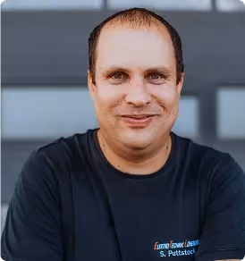 Ein Mann in einem schwarzen T-Shirt lächelt frontal in die Kamera vor einem grauen Hintergrund.