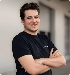 Junger Mann mit schwarzem T-Shirt lächelt und verschränkt die Arme vor einem unscharfen Hintergrund.