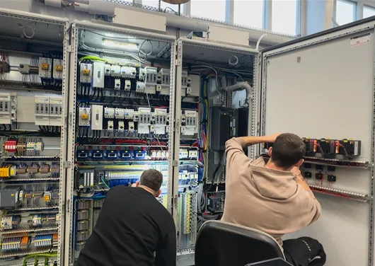 Zwei Techniker arbeiten in einem Schaltschrank mit vielen elektrischen Steuerungen und Kabeln.