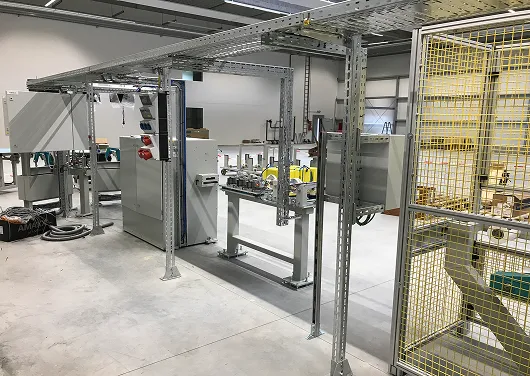Industrielle Fertigungsanlage mit Metallgestell, Schaltschränken und gelbem Schutzzaun in Werkhalle.