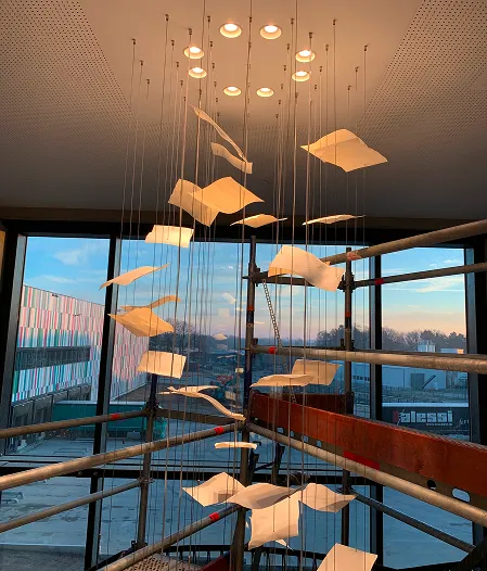Kunstinstallation mit hängenden Papierblättern, durch Fenster sieht man Baugerüst und Stadt bei Sonnenuntergang.