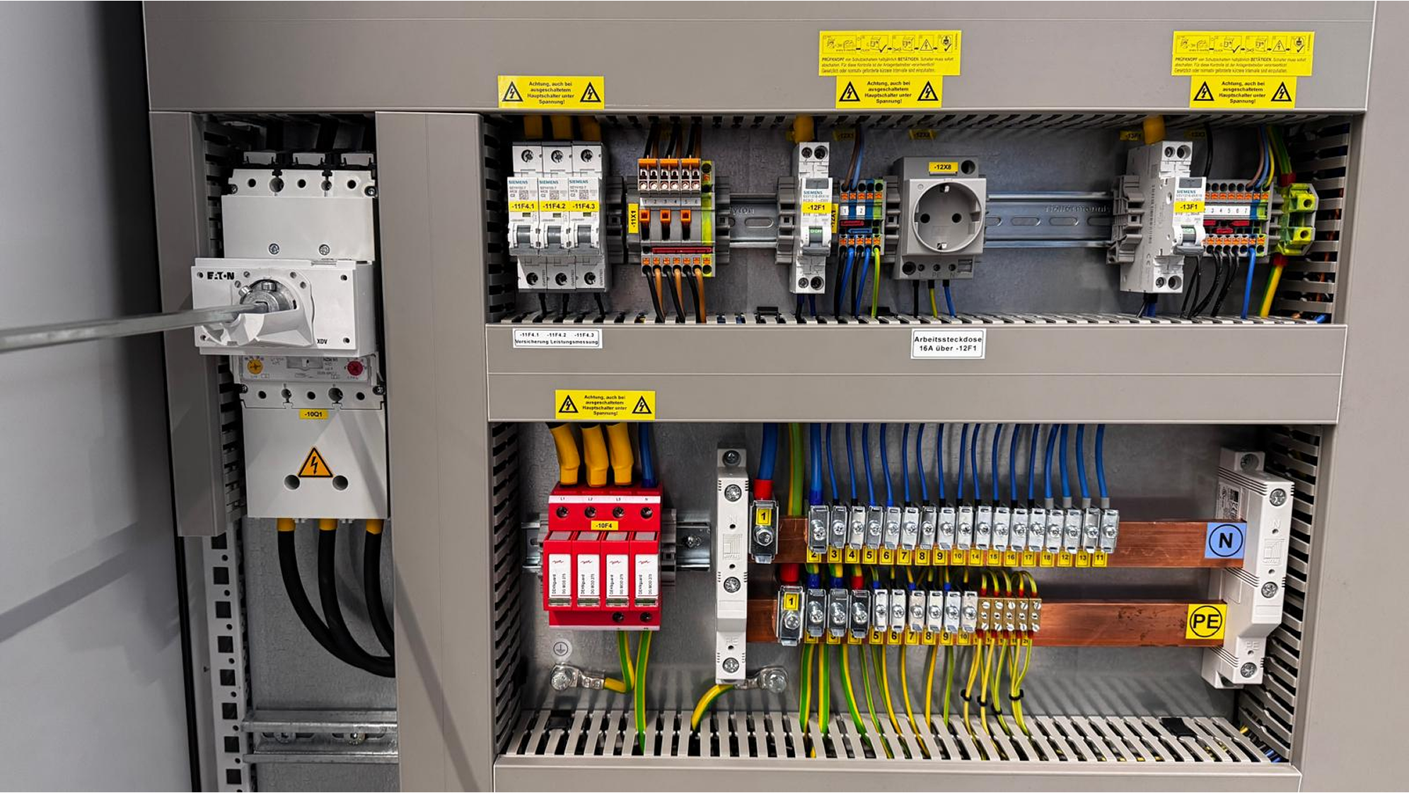 Elektrischer Schaltschrank mit mehreren Sicherungen, Steckdose und geordneten Kabeln in verschiedenen Farben.