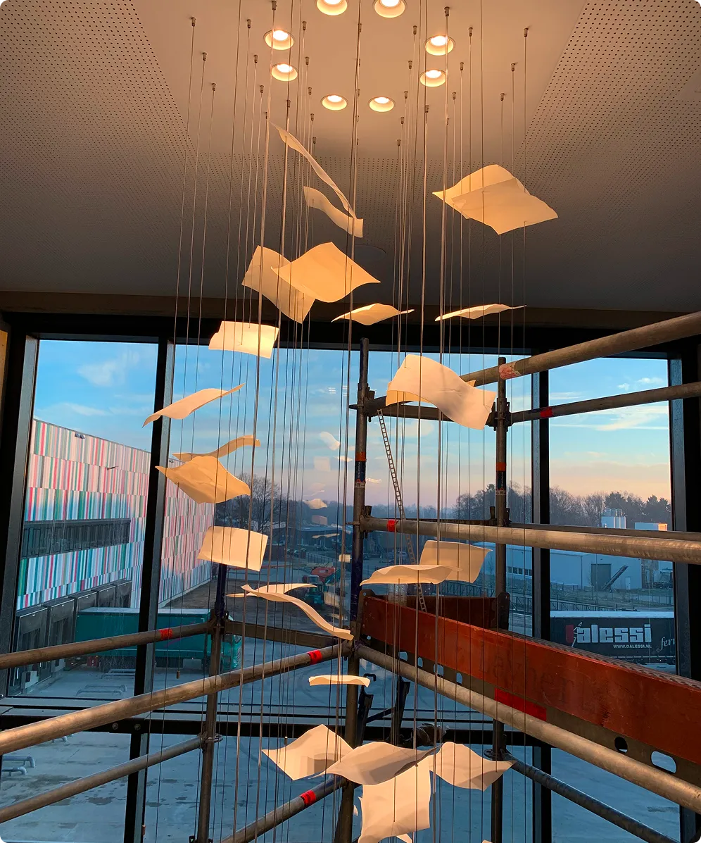 Kunstinstallation mit herabhängenden, leuchtenden Papierbögen in einem Raum mit großen Fenstern.