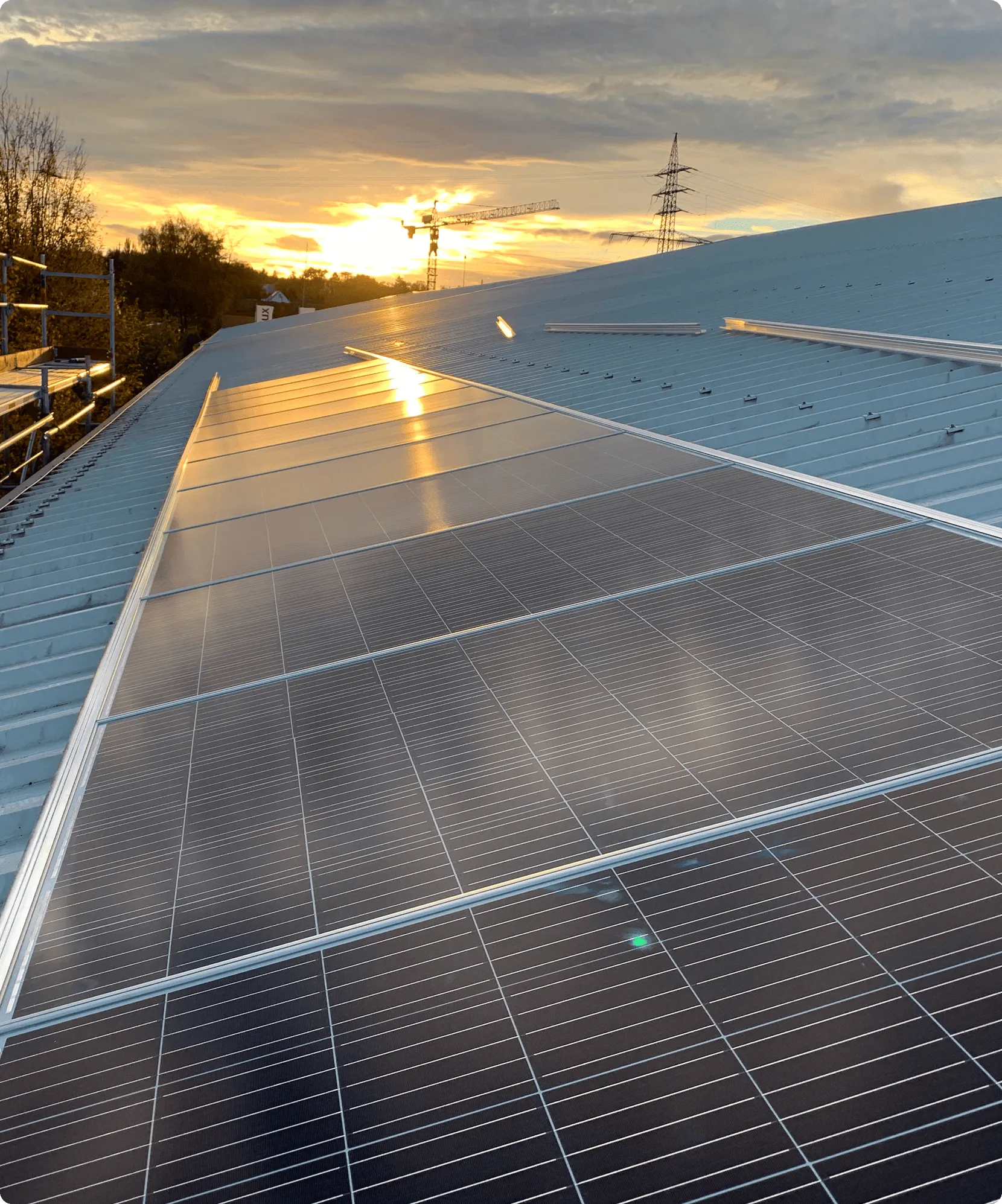Solarmodule auf einem Dach bei Sonnenuntergang mit Baukran und Strommast im Hintergrund.