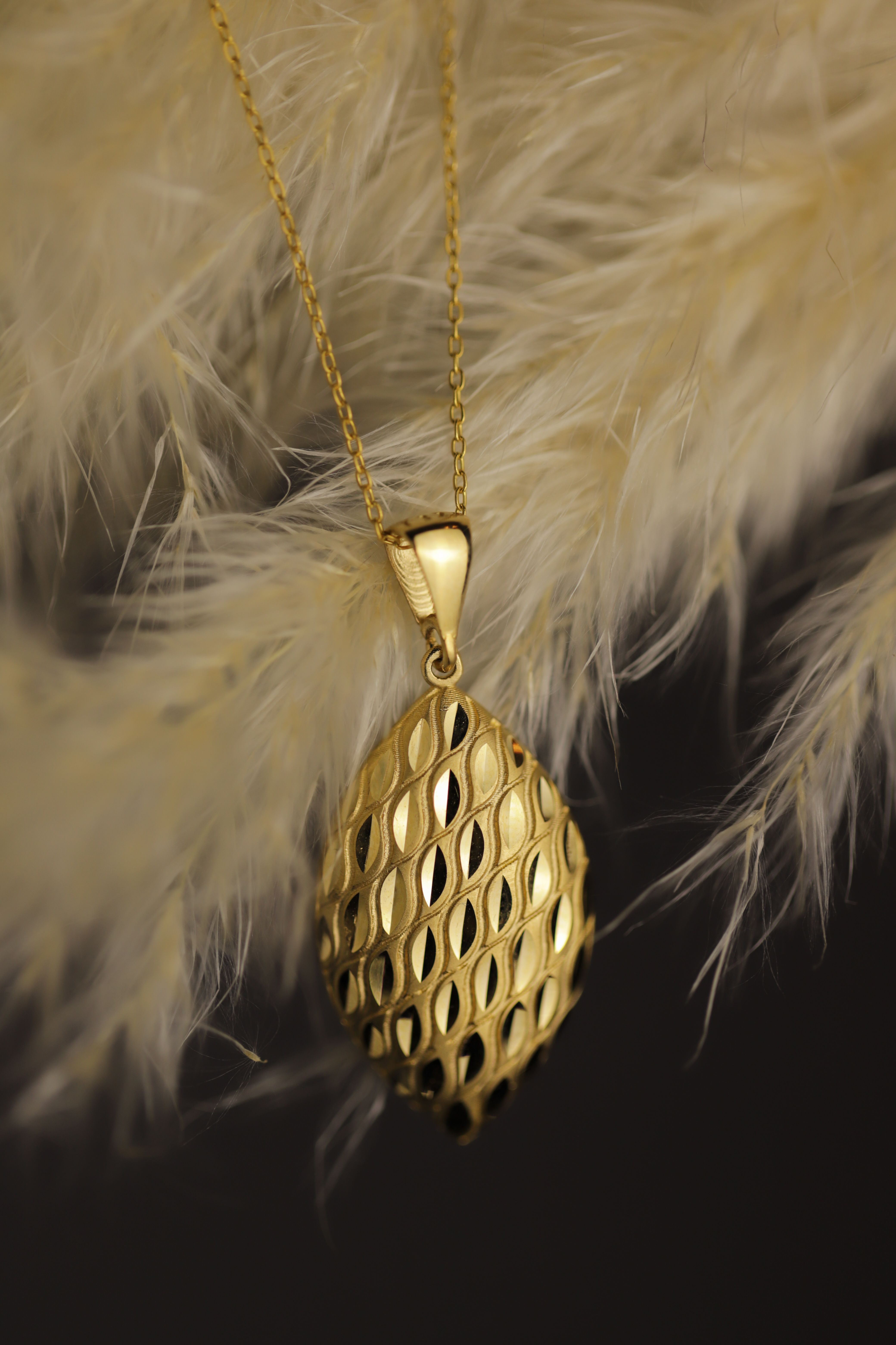 Close-up of a gold pendant with a textured diamond pattern on a gold chain against soft beige feathers.