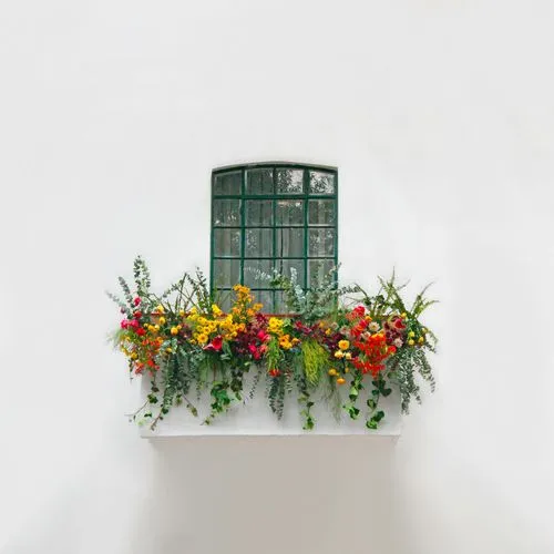 Green window with a flower box filled with vibrant red, yellow, and orange flowers against a white wall.